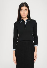 Frau mit langen dunklen Haaren, die einen schwarzen, knöpfbaren Pullover mit hellblauem Kragen und Bündchen sowie einen schwarzen, strukturierten Rock trägt, steht vor einem weißen Hintergrund.