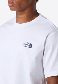 Nærbillede af en person iført en hvid T-shirt med rund hals, der har et lille sort "The North Face" logo på venstre bryst.