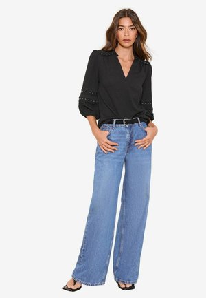 Frau steht mit den Händen in den Taschen, trägt eine schwarze V-Ausschnitt-Bluse mit Puffärmeln und hellblaue Jeans mit weitem Bein sowie schwarze Sandalen.