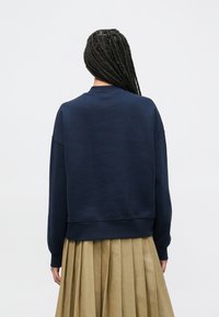 Φούτερ σε σκούρο μπλε ναυτικό χρώμα με άνετη γραμμή, στρογγυλή λαιμόκοψη και μανσέτες με ριμπ ύφανση, συνδυασμένο με πιετέ φούστα σε μπεζ χρώμα. Πλάτη προβολή.