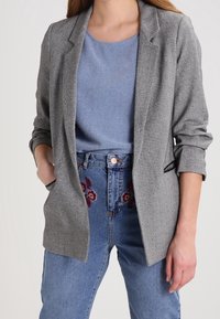 Szary, teksturowany żakiet na jasnoniebieską, błyszczącą bluzkę, połączony z niebieskimi dżinsami z wysokim stanem z kwiatowym haftem.