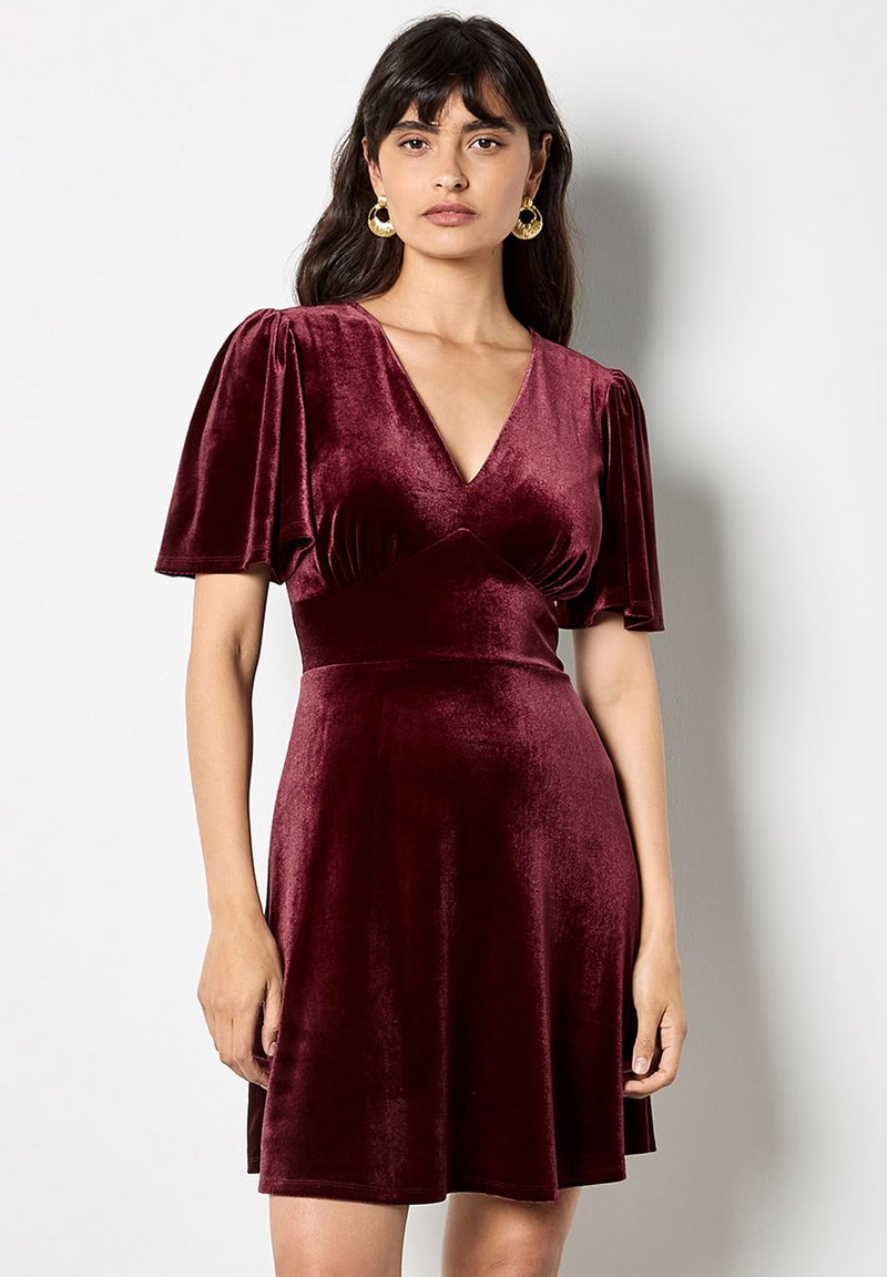 Burgundské sametové šaty s krátkými vystouplými rukávy, hlubokým V-výstřihem a přiléhavým pasem, který se rozšiřuje do stylu skater. Zlaté kruhové náušnice.