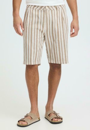 Mann iført beige og hvite vertikale striper shorts til knelengde og beige sandaler med dobbel spenne, stående på en ensfarget bakgrunn.