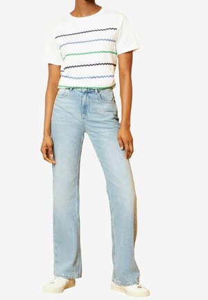 Personne portant un t-shirt blanc avec des rayures ondulées multicolores, un jean droit taille haute bleu clair et des baskets blanches.