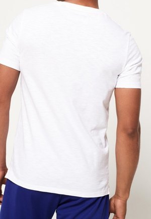 Homme portant un t-shirt blanc uni à manches courtes et un pantalon bleu, vu de dos sur un fond clair.