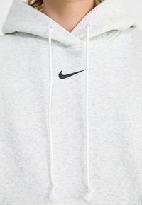 Grijze katoenen hoodie met een trekkoord, voorzien van een zwart Nike-logo op de borst en witte kwastjes aan de trekkoorden. Zachte textuur.
