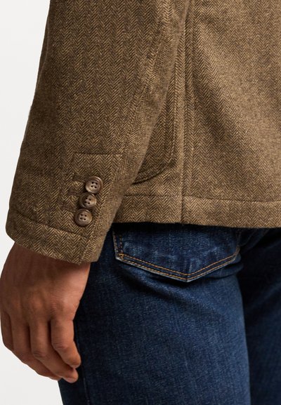 Braune Fischgratjacke mit strukturiertem Finish, ausgestattet mit drei Knöpfen am Ärmel und Seitentaschen, getragen über Jeans.