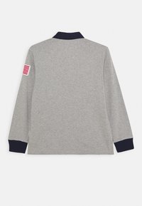 Šedá polokošile s dlouhým rukávem, tmavě modrým límcem a manžetami, s malou červenou a bílou nášivkou na levém rukávu.