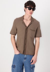 Cămașă scurtă maro cu un design tricotat texturat, cu guler și un buzunar pe piept, purtată cu blugi cargo albaștri.