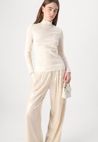 Crèmekleurige fitted top met een hoge gerimpelde kraag, gecombineerd met losse, glanzende, met pailletten bezette beige broek en een kleine beige handtas.