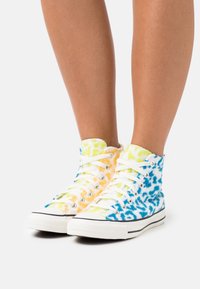 Vysoké tenisky vyrobené z texturované látky v modrých, žlutých a oranžových tie-dye vzorech. Bílé tkaničky a gumové podrážky s černými akcenty.