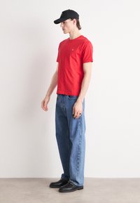 T-shirt en coton rouge, à manches courtes, col rond, associé à un jean bleu en denim ample. Une casquette noire et des chaussures à enfiler complètent l'ensemble.