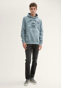 Felpa blu con cappuccio e tasca frontale, con logo e design testuale. Abbinata a jeans scuri e sneaker grigie. Tessuto dalla trama liscia.