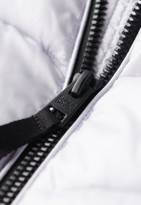 Vit pufferjacka med texturerad yta, utrustad med en svart dragkedja med en logobricka och ett band längs dragkedjan.