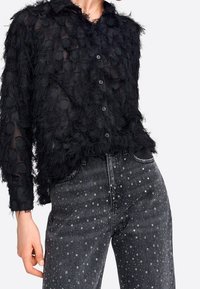 Sort gennemsigtig skjorte med teksturerede, bløde detaljer og knaplukning, parret med mørke jeans med spredte sølvnitter.