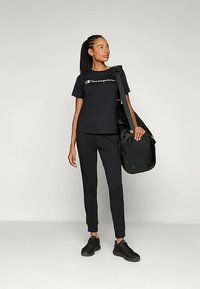 Mustas sportlik riietus, mis koosneb logoga T-sallist, sobivatest pükstest ja mustadest tossudest. Ühel õlal kantakse suurt musta seljakotti.