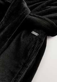 Schwarzer Fleece-Bademantel aus geripptem Stoff, mit gebundenem Gürtel und einem kleinen metallischen Logo-Tag, der die Textur und das schlanke Design betont.