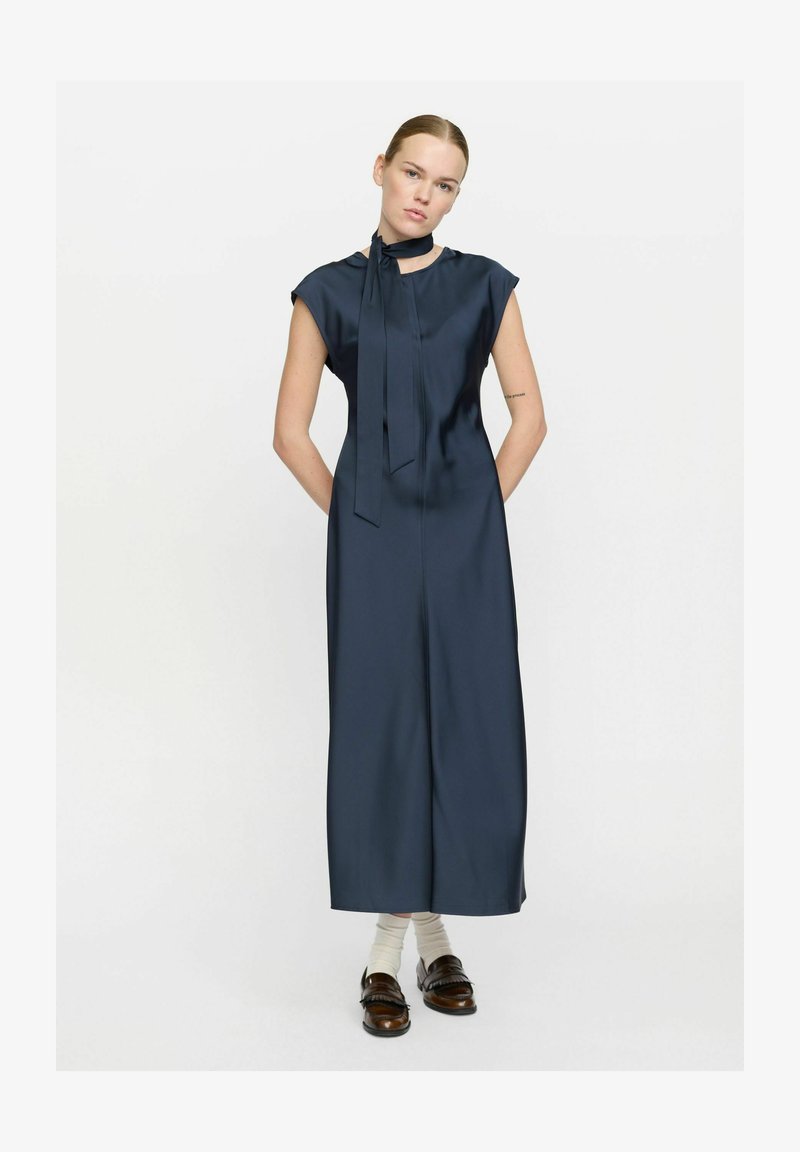 Navyblauwe satijnen jurk met een hoge hals, korte mouwen en een decoratieve strik bij de hals. Het heeft een rechte silhoeet en is volledig lang.