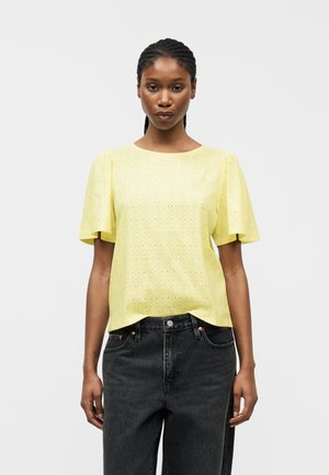 Γυναίκα με πλεξούδες, που φοράει κίτρινη κοντομάνικη μπλούζα με τρύπες και μαύρο τζιν ψηλής μέσης, στέκεται μπροστά σε απλό λευκό φόντο.