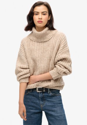Pull oversized beige avec une texture côtelée et un large col roulé, porté sur un jean bleu taille haute et une ceinture noire avec des accessoires en argent.