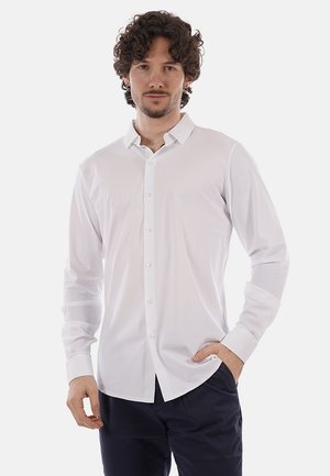 Uomo con capelli ricci che indossa una camicia bianca a maniche lunghe con bottoni e pantaloni scuri, in piedi con una mano in tasca.