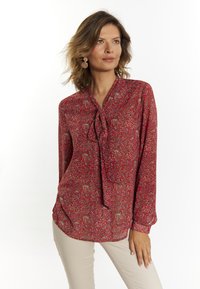 Blusa rossa a maniche lunghe con colletto a fiocco, caratterizzata da un motivo floreale in beige e blu. Realizzata in tessuto leggero e morbido.