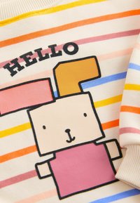 Csíkos pulóver krém színben, több színű csíkokkal. Egy rajzfilmszerű nyuszi mintázattal rendelkezik, rózsaszín testtel és barna fülekkel. A felirat: "HELLO."