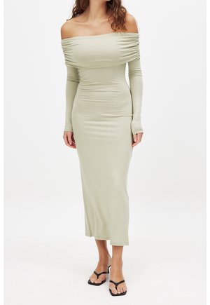 Femme portant une robe midi ajustée à manches longues décolletée couleur sauge pâle, avec des sandales à talons noires ouvertes aux orteils, debout devant un fond blanc uni.