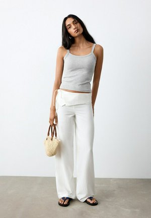 Donna con canottiera grigia, pantaloni bianchi a gamba larga con vita annodata, che tiene una piccola borsa intrecciata, in piedi su un pavimento grigio davanti a una parete bianca.