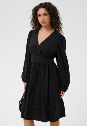 Femme aux cheveux bouclés portant une robe noire à manches longues, coupe évasée, avec un profond décolleté en V et tenant un sac à main noir.