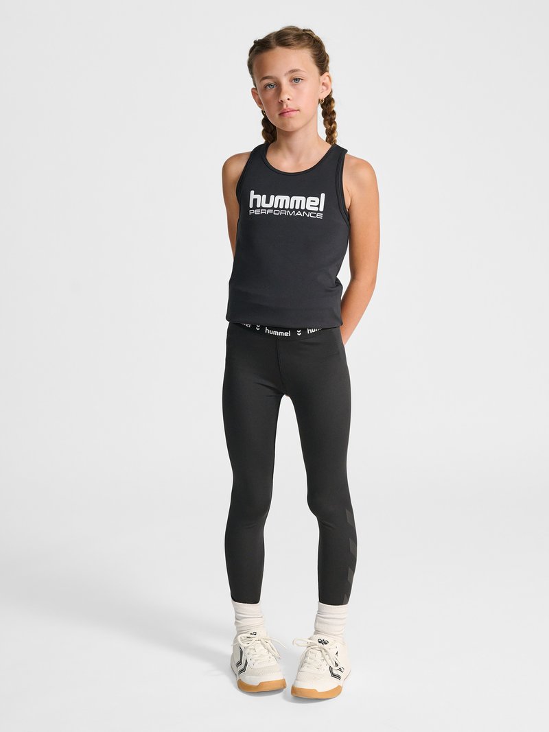 Junges Mädchen mit geflochtenem Haar steht in einem schwarzen „hummel performance“ Tanktop, schwarzen Leggings, weißen Socken und weißen Sneakers vor schlichtem Hintergrund.