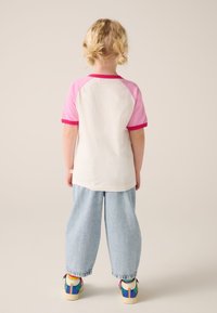 Bērnu T-krekls ar baltu korpusu un rozā raglana piedurknēm, kopā ar plašām gaiši zilām džinsa biksēm un krāsainām sporta kurpēm.