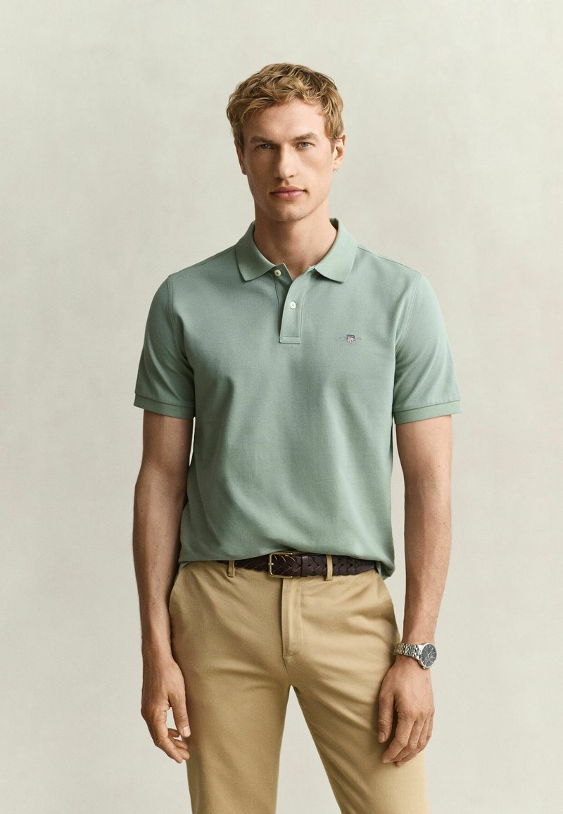 Homme aux cheveux clairs portant un polo vert, un pantalon beige, une ceinture marron et une montre-bracelet argentée sur un fond clair uni.