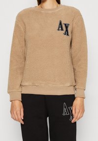 Felpa marrone chiaro in pile con scollo e polsini a coste. Presenta un logo "AX" nero ricamato sul petto e una finitura testurizzata.