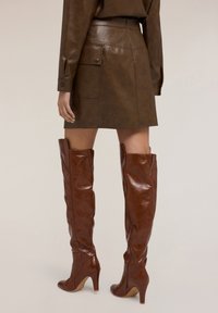 Stivali in pelle marrone alti fino alla coscia con una texture lucida, punta affusolata e tacco sottile alto, dotati di una cerniera laterale per una facile vestibilità.