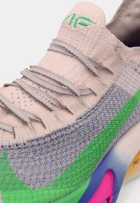 Gros plan d'une basket multicolore avec une tige rose clair, des lacets gris, des détails verts et bleus sur les côtés, et une semelle avec des accents rose et orange.