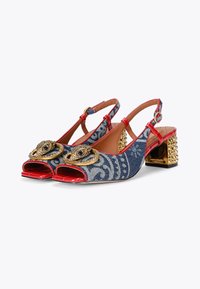 Denim slingback shoes with red patent accents, decorative gold eye motifs, and a textured gold block heel. Features an open toe design.
