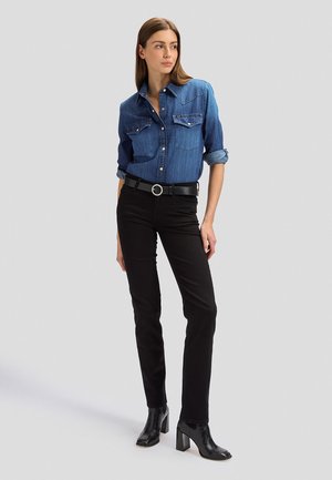 Naine seisab, seljas sinine teksasärk rullitud varrukate ja mustad kitsad püksid, must rihm ümmarguse pandlaga ning mustad kontsaga saapad.