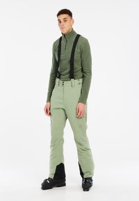 Haut en polaire verte à demi-zip associé à un pantalon de ski vert clair. Les pantalons présentent des bretelles noires et des accents foncés au niveau des chevilles. Bottes de ski noires.