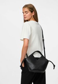 Moteris, dėvinti baltą trumpą rankovėmis marškinius ir juodas kelnes, per petį nešanti struktūrinį juodą odinį rankinį, žiūri žemyn.