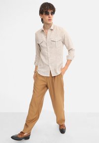 Camicia lunga a maniche lunghe beige con motivo e due tasche a patta, abbinata a pantaloni marroni a vestibilità ampia e scarpe marroni con dettagli neri.