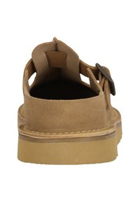 Braune Wildleder-Schuhe ohne Ferse mit flacher Sohle, runder Zehenpartie und seitlichen Ausschnitten. Ein metallischer Schnallenakzent auf einer Seite.