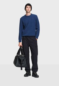 Maglione blu a maglia con scollatura rotonda, pantaloni cargo neri, stivali di gomma neri e borsa a tracolla nera con dettagli sulla tracolla.