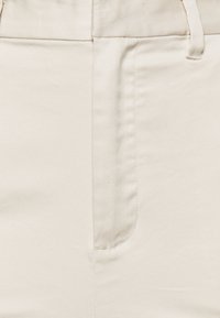 Gros plan sur un tissu couleur crème sur un pantalon, montrant une poche latérale, une passant de ceinture et des détails de couture visibles.