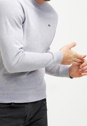 Mężczyzna w jasnoszarej bluzie z małym zielonym logo krokodyla, klaskający w ręce, w czarnych spodniach, na tle białym.