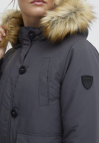 Grå pufferjakke med faux fur hette. Har knappelukking, sidelommer og et svart logo-patch på armen. Glatt tekstur.