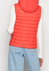 Bageste udsigt af en person iført en rød vatteret hættetrøje over en hvid ribbet langærmet bluse og lyseblå denim jeans.