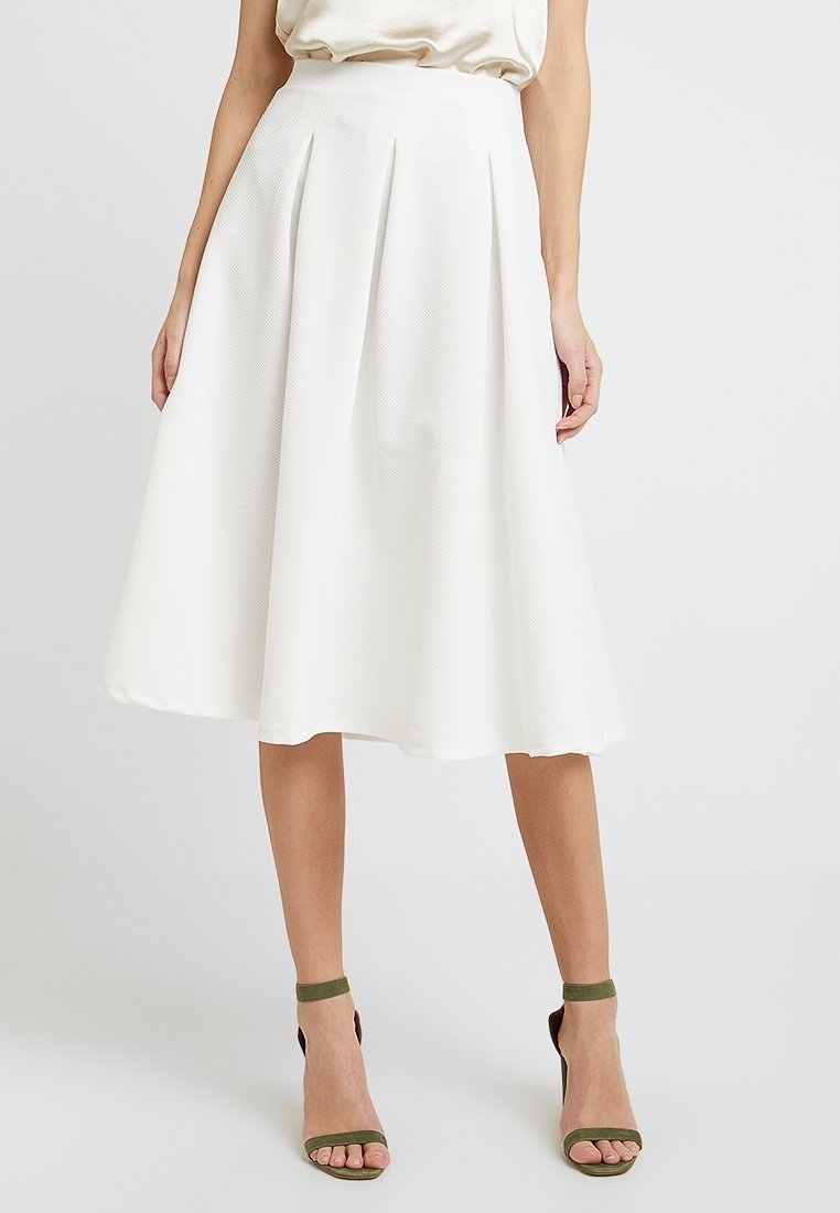 Femme portant une jupe midi blanche taille haute en ligne A avec des plis, associée à un chemisier crème et des escarpins à brides à la cheville verts olive.