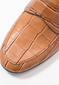 Tan crocodile-pattern leather shoe with a rounded toe, featuring a stitched detail and a strap across the upper. Smooth texture.