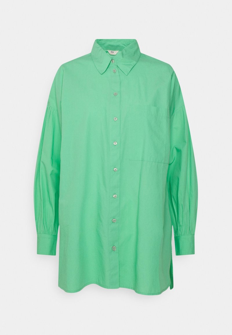 Chemise ample à manches longues de couleur verte, boutonnée, avec col pointu et poche poitrine, présentée sur un fond blanc.